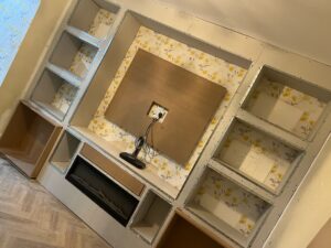 Modern living room with a built-in entertainment center featuring gray shelving, a sleek wall-mounted TV area, and floral wallpaper accents. The design includes space for decorative items and a fireplace, creating a stylish focal point for the room.