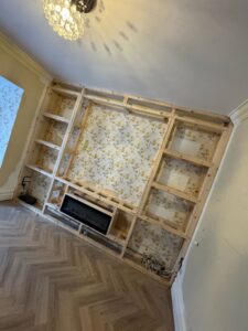 Modern living room featuring a custom-built wooden shelving unit with a floral wallpaper backdrop, designed for storage and display. The room has a light color scheme and wooden flooring, creating a warm and inviting atmosphere.