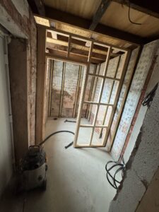 Interior view of a construction site showing exposed wooden framing and a partially installed door frame, with construction tools and equipment visible on the floor.
