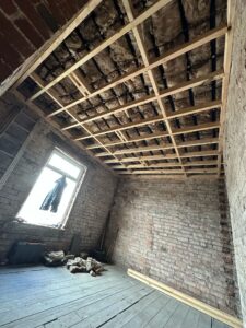 Interior view of a partially constructed room featuring exposed brick walls, a wooden ceiling frame, and a window with a coat hanging nearby. The floor is bare, indicating ongoing renovation work.