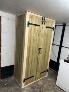 Wooden storage cabinet with double doors and black metal hardware, positioned in a basement setting. The cabinet features a natural wood finish and provides ample space for organization.