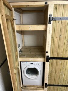 Compact laundry storage solution featuring a washing machine on the bottom shelf and two wooden shelves above for organization. The design includes wooden paneling and sturdy hinges, ideal for small spaces.