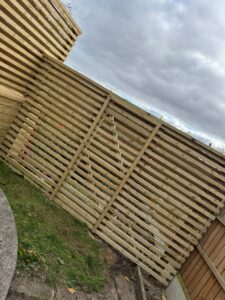 Wooden slatted fence panels installed in a garden, with a cloudy sky in the background, showcasing a blend of natural materials and outdoor design elements.