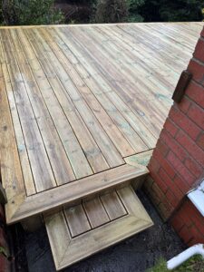 Wooden deck with steps leading to a brick wall, showcasing a well-maintained outdoor space. The deck features a smooth surface with a natural finish, surrounded by greenery. Ideal for enhancing backyard aesthetics and functionality.