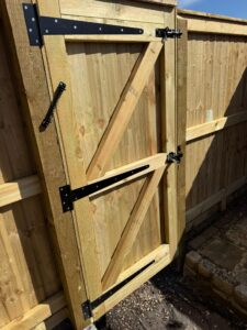 Wooden garden gate with black metal hinges and latch, set against a wooden fence, showcasing craftsmanship and durability in outdoor security solutions.