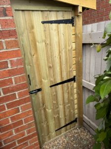 Wooden garden gate with black metal hinges and latches, positioned next to a brick wall and surrounded by gravel and greenery. The gate features a natural wood finish and a secure design, ideal for outdoor spaces.