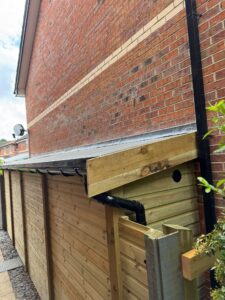 Side view of a wooden garden shed with a sloped roof, featuring a rainwater gutter and drainage pipe, adjacent to a brick wall. The shed is constructed with vertical wooden panels and is surrounded by gravel and greenery.