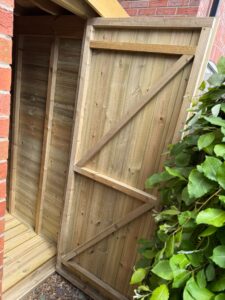 Wooden shed door partially open, revealing a spacious interior with wooden paneling and flooring, situated next to a red brick wall and surrounded by lush green foliage. Ideal for storage solutions and garden organization.