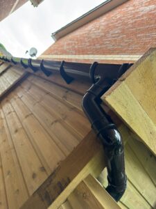 Close-up view of a black gutter system attached to a wooden structure, showcasing the curved downspout and brackets against a backdrop of brick walls and cloudy sky.