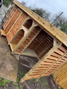 Wooden garden shed under construction, featuring a slanted roof, wooden slats, and a door, set on a gravel and grass area. The structure is partially completed, showcasing craftsmanship and design suitable for outdoor storage.