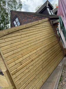 Tilted wooden fence panel leaning against a brick house, with trees and clothes drying in the background, showcasing potential storm damage or structural instability.