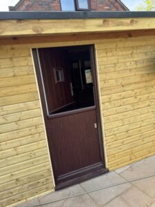 Wooden exterior of a garden shed featuring a brown door, with a glimpse of interior space visible through the doorway. The structure has a rustic design, complemented by a stone patio.