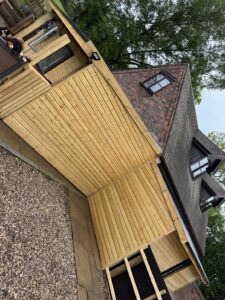 Modern wooden house exterior with a slanted roof, featuring a combination of wooden cladding and brick. The image showcases a patio area with a gravel pathway, surrounded by trees. Two dogs are seen lounging on the deck, adding a lively touch to the serene outdoor setting.