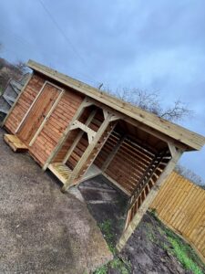 Wooden garden shed with a sloped roof, featuring a door and an open side, set against a cloudy sky. The shed is constructed from timber and includes a small wooden platform in front. Surrounding landscape shows gravel and patches of grass, with a wooden fence in the background.