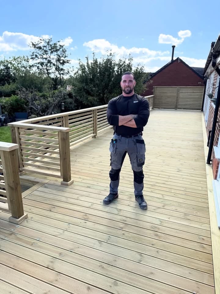 Professional carpenter standing on a newly constructed wooden deck, showcasing craftsmanship and attention to detail, with a clear blue sky and greenery in the background.