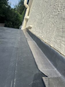 Flat roof edge with black waterproofing membrane, showing detail of the transition to the textured wall surface and drainage pipes, surrounded by greenery.