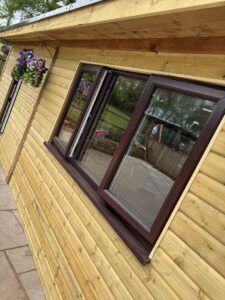 Exterior view of a wooden cabin featuring large brown windows and a hanging flower basket, showcasing the natural wood texture of the walls and a well-maintained outdoor area.