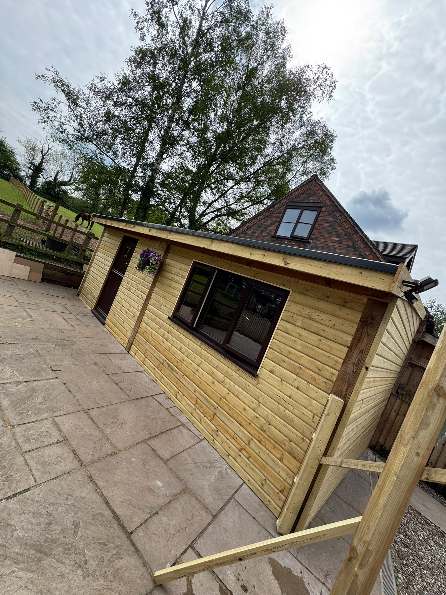 Wooden garden shed with a sloped roof, large windows, and a stone patio, surrounded by trees and a grassy area. The structure features natural wood siding and a decorative hanging plant.